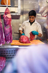 Hispanischer männlicher Metzger steht hinter einer Metalltheke und schneidet frisches Fleisch während der Arbeit auf dem Markt - ADSF36669