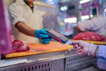 Weichzeichner eines anonymen Verkäufers in Handschuhen, der einer Kundin beim Verkauf von Fleisch auf dem Markt ein Terminal mit Kreditkarte übergibt - ADSF36670