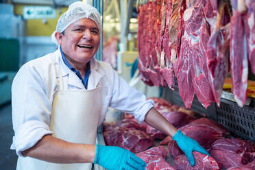 Älterer Mann in Uniform berührt frisches Fleisch am Verkaufsstand bei der Arbeit in der Metzgerei auf dem Markt - ADSF36672