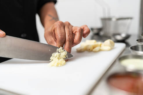 Crop anonymous chef cutting garlic with sharp knife on chopping board while cooking in kitchen of restaurant against blurred background - ADSF36678