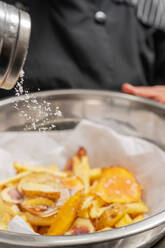 Anonymer Koch in schwarzer Uniform, der Salz in eine Glasschüssel mit Bratkartoffeln streut, während er in einer modernen hellen Küche kocht - ADSF36681