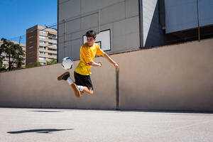Junger männlicher Spieler, der beim Üben Fußball gegen ein Gebäude tritt - ADSF36685