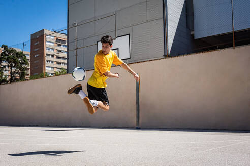 Young male player kicking football while practicing soccer against building - ADSF36685