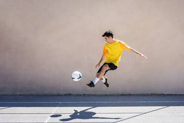 Junger männlicher Spieler, der beim Üben Fußball gegen ein Gebäude tritt - ADSF36686