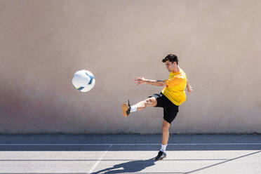 Junger männlicher Spieler, der beim Üben Fußball gegen ein Gebäude tritt - ADSF36687