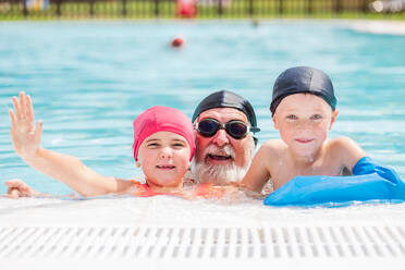 Aufgeregte kleine Kinder mit Mützen, die strahlend lächeln und in die Kamera schauen, während sie mit einem glücklichen bärtigen Großvater in den Sommerferien im Freibad schwimmen - ADSF36693