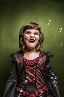 Adorable kleines Mädchen mit gruseligen Make-up im Kostüm spielen mit Seifenblasen und suchen weg gegen bunten Hintergrund während Halloween-Party - ADSF36706