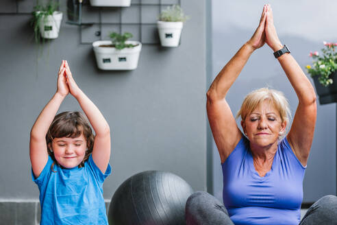 Ältere Frau und kleines Mädchen mit geschlossenen Augen und erhobenen Armen beim Meditieren zu Hause - ADSF36719