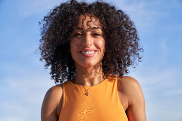 Cheerful Hispanic female with curly hair looking at camera while standing against blue sky on sunny summer day - ADSF36738