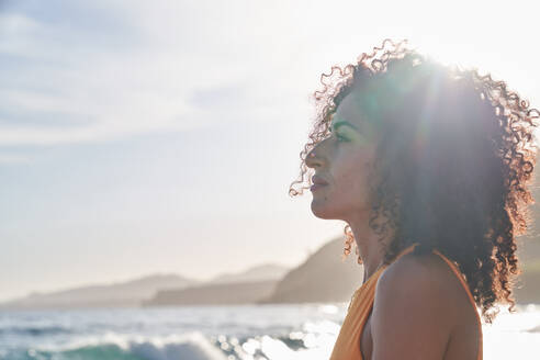 Hispanische Frau mit lockigem Haar, die am Meer steht und wegschaut, an einem sonnigen Tag - ADSF36739