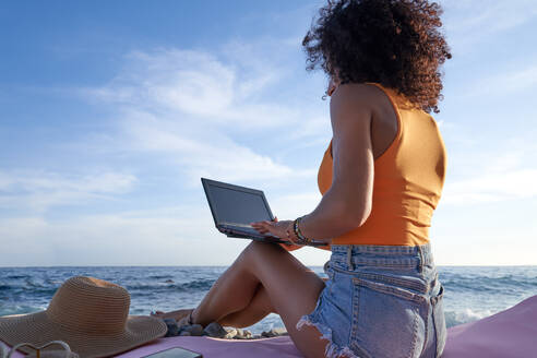 Seitenansicht einer Frau, die auf einem Netbook im Internet surft, während sie an einem Sommertag in einer Küstenregion am Meer sitzt - ADSF36740