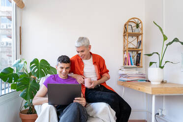 Lächelndes schwules Paar mit Laptop auf einem Sessel zu Hause - ADSF36754
