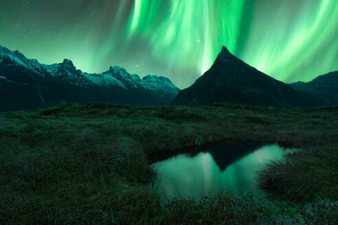 Runder, von Gras umgebener See in der Nähe hoher, rauer, schneebedeckter Felsklippen unter hellen Polarlichtern auf den Lofoten - ADSF36814