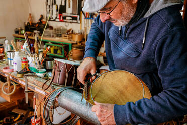 Männlicher Handwerker, der eine Schere benutzt, während er die Metallkanten einer Trommel abschneidet, während er in einer professionellen Lichtwerkstatt arbeitet - ADSF36833