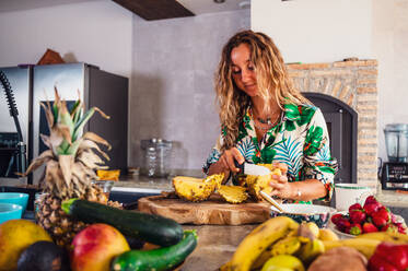 Focused female cutting ripe pineapple on wooden chopping board near various fruits and berries while standing in light modern kitchen - ADSF36844