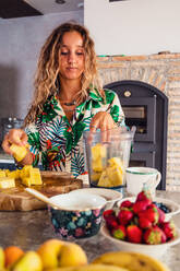 Fokussierte Frau, die frische gehackte Ananas in den Mixer gibt, während sie am Tisch mit verschiedenen gesunden Lebensmitteln in der hellen Küche kocht - ADSF36846