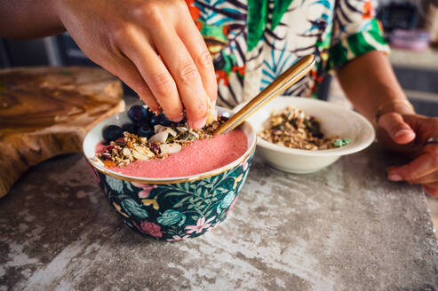 Crop anonyme weibliche Putting knackig süßen Granola in leckeren rosa Smoothie Schüssel mit Blaubeeren während des Frühstücks in der Küche zu Hause - ADSF36852