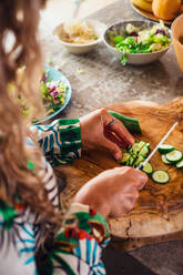 From above of crop anonymous female cook cutting fresh green cucumber on chopping board while cooking salad in light kitchen - ADSF36858