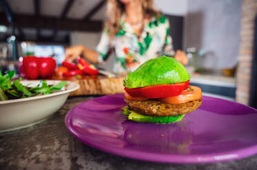 Weichzeichner von Avocadohälften, die einen Burger mit Gemüse und veganer Frikadelle auf einem Teller neben einer Frau in der Küche zu Hause bilden - ADSF36869