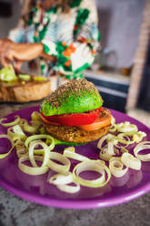Weichzeichner von Avocadohälften, die einen Burger mit Gemüse und veganer Frikadelle auf einem Teller neben einer Frau in der Küche zu Hause bilden - ADSF36871