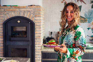 Happy blond female carrying plate with healthy vegetarian burger and looking at camera with smile during lunch in light kitchen at home - ADSF36872