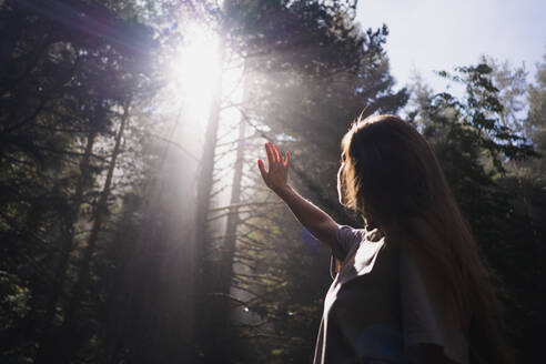 Von unten Seitenansicht einer weiblichen Reisenden, die mit ausgestrecktem Arm im Wald steht und sich vor den hellen Sonnenstrahlen versteckt, die durch die Äste dringen und wegschauen - ADSF36876