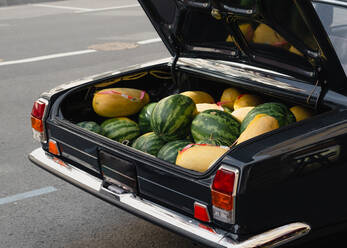 Hoher Winkel von reifen Melonen und Wassermelonen für den Verkauf in geöffneten Kofferraum von Vintage schwarzes Fahrzeug auf Asphalt Straße geparkt auf Stadtstraße platziert - ADSF36903