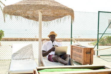 Full body of African American businessman with netbook surfing cellphone while lying on lounger under parasol in coastal area on summer day - ADSF36927