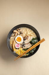 From above of appetizing Japanese ramen with boiled egg and mushrooms served in bowl with wooden chopsticks against beige background - ADSF36960