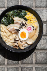 Draufsicht auf appetitliche japanische Ramen mit Pilzen und Ei, serviert auf einem gepflasterten Bürgersteig in einer sonnigen Straße in der Stadt - ADSF36966