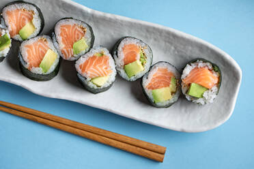 From above set of tasty Futomaki Norwegian sushi rolls with salmon and avocado served near chopsticks on blue background in light studio - ADSF36972