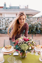 Positive Frau in Kleidern arrangiert Tisch mit frischen Blumen und leckeren Gerichten auf der Terrasse eines Gebäudes an einem Sommertag - ADSF37001