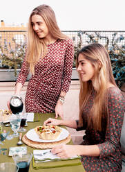 Zufriedene Freundinnen in Kleidern beim Abendessen am Tisch mit Gläsern und leckerem Salat auf der Terrasse im Sommer - ADSF37006