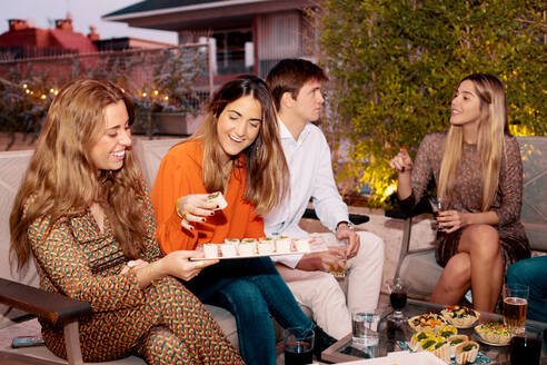 Group of smiling friends chatting and having dinner with tasty appetizers while sitting at table on terrace in evening time - ADSF37014