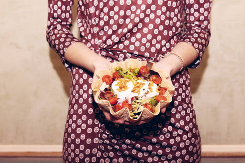 Cropped unrecognizable female with long hair in dress looking away while holding salad in tortilla bowl near wall during dinner in light modern restaurant - ADSF37020