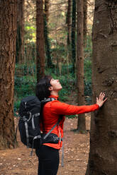 Seitenansicht einer Frau mit Rucksack, die einen Baumstamm berührt und nach oben schaut, während sie den Wald an einem Sommertag erkundet - ADSF37043