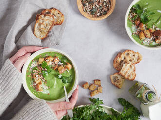 Top view of crop woman holding tasty green cream soup with parsley topped with croutons placed on table with greens in light kitchen - ADSF37069