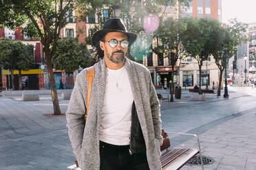 Erwachsener hispanischer Mann in modischer Kleidung mit Sonnenbrille und Hut, der die Hände in den Taschen hält und wegschaut, während er in der Nähe einer Bank auf einer sonnenbeschienenen Stadtstraße steht - ADSF37077