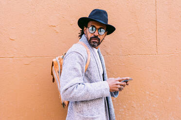 Hispanic male in stylish clothes with hat and sunglasses leaning on wall and browsing cellphone while standing on pavement on city street - ADSF37079