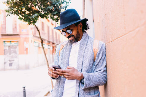 Hispanischer Mann in stilvoller Kleidung mit Hut und Sonnenbrille, der sich an eine Wand lehnt und in seinem Handy blättert, während er auf dem Bürgersteig einer städtischen Straße steht - ADSF37080