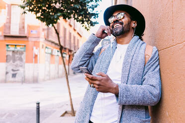 Adult Hispanic man in trendy clothes with hat and sunglasses putting on TWS earphones and browsing playlist on smartphone while resting on city street in daytime - ADSF37081