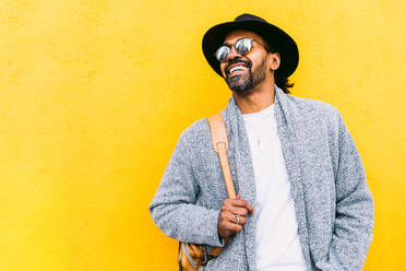 Fröhlicher hispanischer Mann in stilvoller Kleidung mit Tasche auf der Schulter, der wegschaut und lacht, während er an einer leuchtend gelben Wand auf einer Stadtstraße steht - ADSF37083