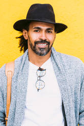 Fröhlicher hispanischer Mann in stilvoller Kleidung mit Tasche auf der Schulter, der in die Kamera schaut und lacht, während er an einer leuchtend gelben Wand auf einer Stadtstraße steht - ADSF37086