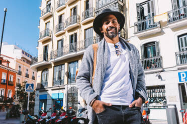 Adult Hispanic male in trendy clothes with sunglasses and hat holding hands in pocket and looking away while standing on street - ADSF37088