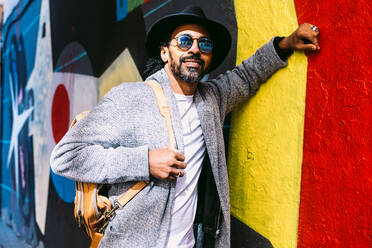 Positive Hispanic male in trendy clothes with backpack leaning on colorful graffiti wall and looking at camera with smile on sunny day on city street - ADSF37092