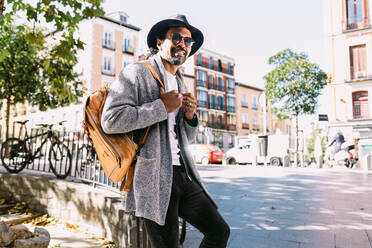 Hispanischer Mann in modischer Kleidung mit Rucksack, der lächelt und wegschaut, während er sich an einen Zaun lehnt und auf einer sonnenbeschienenen Straße ausruht - ADSF37097