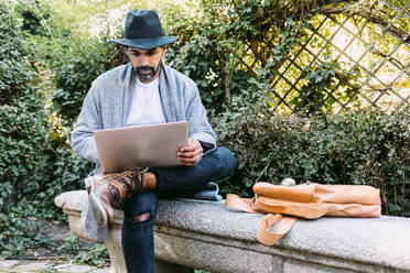 Adult Hispanic man in trendy clothes and hat browsing netbook while sitting on stone bench and working remotely from park - ADSF37100