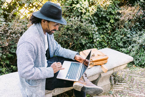 Erwachsener hispanischer Mann in trendiger Kleidung und Hut, der auf einer Steinbank sitzend auf seinem Netbook surft und aus der Ferne vom Park aus arbeitet - ADSF37101