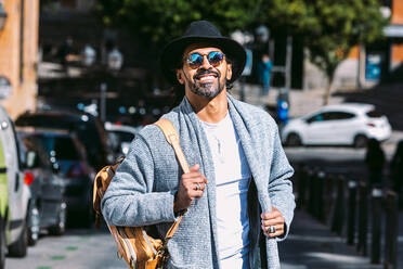 Erwachsener Hispanoamerikaner in modischer Kleidung mit Sonnenbrille und Hut, der die Hände in den Taschen hält und in die Kamera schaut, während er auf der Straße steht - ADSF37103