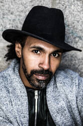 Serious bearded Hispanic male in stylish clothes and hat looking at camera against stone wall on street - ADSF37104